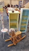 View of children's coat rack with heart-shaped hooks, polka-dot stool at base, yellow rolling shelf unit, blue bookcase, wooden rocking horse, and small night light on shelf top