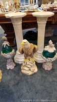 Two white plaster pedestals, eagle sculpture, two seashell compotes with green glass egg and bowl displayed on carpeted floor.