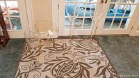 Two very tall clear plastic stemware glasses and one smaller clear plastic stemware glass standing on a decorative indoor-outdoor rug in front of a glass door.