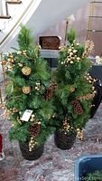 Pair of matching artificial green pine trees with gold decorations including spherical ornaments, berries, pine cones, and placed in decorative dark pots under a stairway.