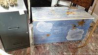 View of gray metal 2-drawer file cabinet and painted wooden toy chest with sky/cloud and childlike design.