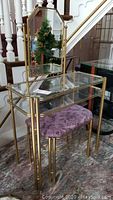 Full view of glass and brass vanity table with attached mirror and purple upholstered seat below.
