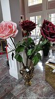 Three giant artificial roses in a brass-colored urn planter with cupid motifs. One rose is light pink, two are dark red. Pine green leaves and pussy willow reeds accompany the flowers. Arranged on a marble floor in a well-lit interior.