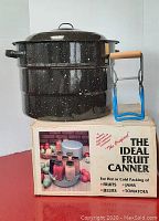 The Ideal Fruit Canner 21-quart enamel canning pot on original box next to jar lifter with blue rubber handles and wooden grip.