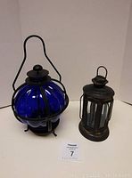 Two patio lanterns side by side on a surface, showing the blue glass lantern and the black metal lantern with a rod iron handle.