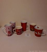 Six holiday Starbucks mugs arranged in a single row, showing different colors and designs including pink Christmas, red Christmas with deer, and red Christmas with bird.
