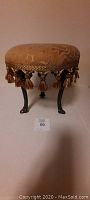 Front view of round cloth covered footstool with iron legs and tassel fringe, shows fabric pattern and detail of legs.