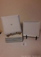 Stack of 12 white square dinner plates with one propped vertically to show shape and edges. Plates have smooth surfaces with a subtle raised lip.