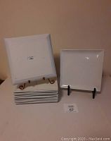 Photo showing 12 stacked white square plates with one plate displayed upright on a stand, showing smooth surface and shape.