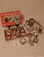 Clear view of 14 vintage metal cookie cutters arranged beside a boxed set of 12 Barnyard cookie cutters on a table, showing the shapes and box design.