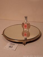 Round beveled glass footed mirror tray with a single bottle of pink-labeled cologne. The bottle is placed on the tray, showing reflection.