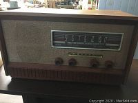 Front view of Marconi tube radio showing dial with AM frequencies and four control knobs, beige fabric speaker grille, and wooden cabinet.