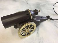 Side view of the lamp showing the cannon shape and gold-tone wheels with power cord attached.