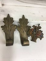 Three vintage metal curtain tie back holders: two brass-colored leaf designs and one painted floral basket design.