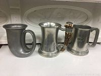 Lot of three vintage pewter mugs with distinct shapes and finishes.
