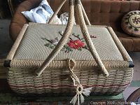 Photo showing woven sewing basket with two handles and floral embroidery on the lid, placed on a surface with couch background.