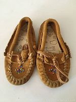 Pair of tan suede moccasins side by side, showing detailed beadwork and braided leather laces.