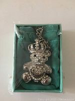 Photo showing silver-tone Juicy Couture teddy bear necklace pendant encrusted with rhinestones and crown detail, inside original turquoise box with clear plastic wrapping.