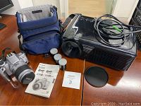 Overview of camera, case, manual, film rolls, projector, lens cap and cable on table