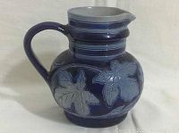 Side view of a deep blue glazed pottery jug creamer with embossed leaf and grape design, curved handle and spout.