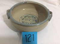 Front angle view of the handcrafted pottery bowl showing the beige glaze, blue rim, and side handles with interior circular design.