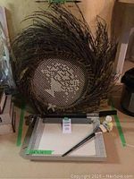 Two brown twig wreaths stacked vertically, 24 inches diameter each. Front wreath has decorative mesh center with textured pattern. Below wreaths is a 16 x 12 inch paper cutter with metal base and clear ruler, plus small tube of art masking fluid and brush.