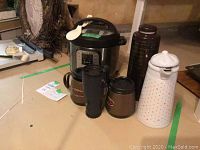 Photo showing the 6 quart Instant Pot with digital control panel and five assorted coffee thermoses including one white ceramic with red dots, one tall brown plastic thermos, and three insulated mugs with lids.