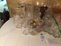 Clear glass kitchenware including jars, pitchers, vases, and glass covered pie stand arranged on a white table