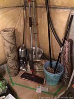 Photo showing two vacuums, wet mop, two rolled doormats, and baskets arranged in basement or storage area.