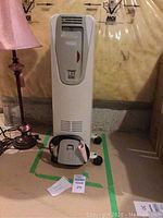 Full front view of the white DeLonghi heater placed on a concrete floor with surrounding items including a pink lamp and cords.