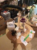 Photo of a round wooden table displaying a boudoir hat lamp and various decorative items including porcelain figurines, trinket boxes, a white pitcher, and other small decor pieces.