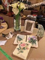 View of the decorative items including floral plaques, picture frame, papier-mâché cat, glass bird figurine, and tall vase with calla lilies on a table.
