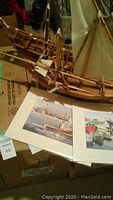 Two wooden sailboats in foreground, open book displaying two signed prints with maritime scenes