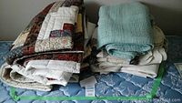 Stack of multiple quilts and blankets including patchwork quilts and a turquoise knitted blanket folded on twin mattress.