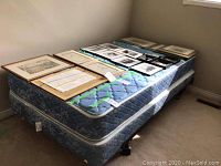 Single bed mattress and box spring stacked, with framed pictures on top around the edges of the mattress, showing quilted blue pattern.