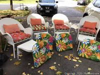 Four white wicker chairs each with seat and back cushions, accompanied by four matching white wicker foot stools. Outdoor setting with fallen leaves on blacktop.