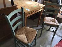 Photo showing two green ladder-back chairs with woven seats next to wooden table.