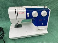 Front view of white and blue plastic Singer sewing machine showing needle area, brand name, and sewing platform.