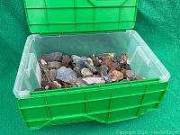 Green plastic bin filled with various rough mineral and rock specimens in assorted colors and stages of processing.