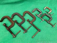 Seven steel C-clamps laid out on green fabric showing varying sizes and wear.