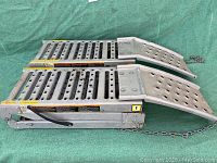 Pair of folded metal loading ramps with perforated surface, safety chains, and locking mechanisms laying on green surface.