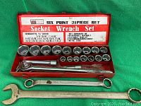 Open metal case showing 16 six-point sockets, reversible ratchet, flexible bar, and extension bars, along with two oversized combination wrenches outside the case.