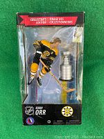 Front view of Bobby Orr figure in Boston Bruins uniform, packaged with Stanley Cup trophy replica in window display box.