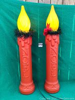 Pair of red plastic Christmas candle lighter displays with yellow illuminated flame tops and Noel embossing, decorated with green garland and bow