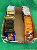 Top view of multiple rows of sealed trading card packs in a cardboard box, showing variety of sports and brands.