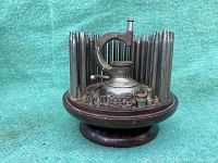 Top and angled views of antique watchmakers staking tool set with numerous metal staking punches arranged vertically around a circular wooden base.