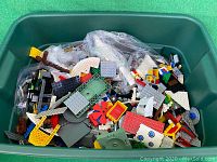 Green plastic bin filled with assorted LEGO bricks, plates, slopes, and minifigure parts