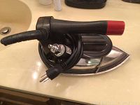 Top and side angled view of Pacific gravity feed steam iron, showing black and red handle with attached power cord and metal soleplate.