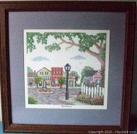 Framed handmade cross stitch artwork 'Our Town' showing entire piece in wooden frame with gray-blue mat.