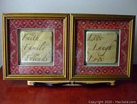 Two plaques with gold frames and red patterned backgrounds 'Faith Family Friends' and 'Live Laugh Love' displayed side by side on wooden stand.